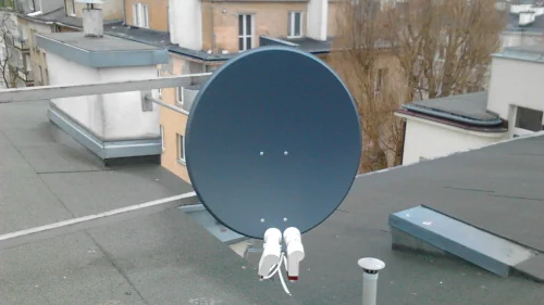 Szara antena satelitarna z dwoma białymi konwerterami zamontowana na dachu, z widokiem na pobliskie budynki i drzewa