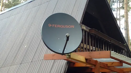Satelitarna antena przy domu Grafitowa antena satelitarna z napisem Ferguson zamontowana na drewnianym wsporniku na zewnątrz domu letniskowego ze skośnym dachem