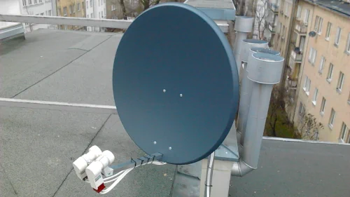 Satelitarna antena na dachu Szara antena satelitarna zamontowana na dachu z podłączonymi do niej kablami, otoczona stalowymi kominami i budynkami na dalszym planie