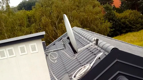 Satelitarna antena na kominie Satelitarna antena zainstalowana na dachu domu z szarym metalowym dachem, w tle widoczne są drzewa i trawiaste pole