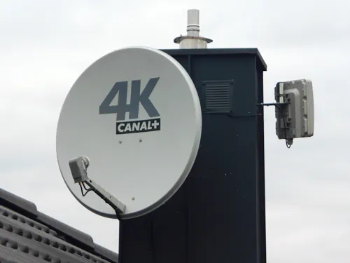 Satelitarna antena Canal+ Antena satelitarna na dachu z logo Canal+ 4K, zamontowana na szarym kominie obok małej anteny internetowej na tle zachmurzonego nieba