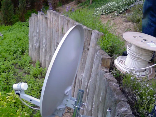 Satelitarna antena w ogrodzie Antena satelitarna zainstalowana w ogrodzie z drewnianym płotem na tle zielonej trawy, obok szpula z kablem koncentrycznym